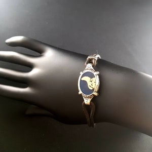 May include: A blue and gold oval charm bracelet with a leather cord. The charm features a stylized design of a bird.