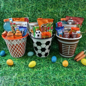 May include: Three themed gift baskets on a green surface. One is a basketball net filled with snacks and orange and white eggs. Another is a soccer ball bucket, and the third is a football-themed bucket. Easter eggs and carrots are scattered around.