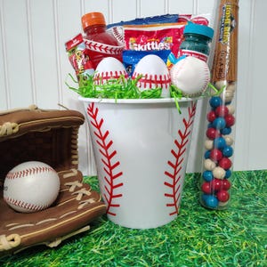 Puede incluir: Una cesta de regalo con temática de béisbol con un cubo blanco con costuras rojas, lleno de bocadillos, huevos de Pascua decorados con béisbol y una pelota de béisbol. También se incluye un guante de béisbol y un tubo de chicles. El fondo es verde.