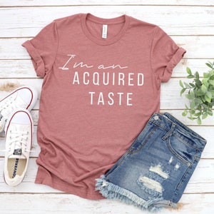 May include: A maroon t-shirt with the text "I'm an acquired taste" in white lettering. The shirt is laid flat on a white wooden surface with a pair of white sneakers, denim shorts, and a small potted plant.