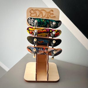 May include: A wooden skateboard display stand holding four miniature skateboards. The top of the stand has the name "EDDIE" cut out. The skateboards have colourful graphics and the words "Tony Hawk" are visible on one.