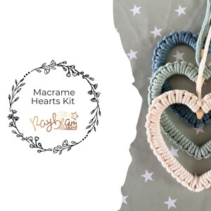 May include: Three macrame hearts in shades of blue and cream are stacked, with a hanging loop. The hearts are on a star-patterned background. The image includes text that reads "Macrame Hearts Kit" and "Raybeam Makes".