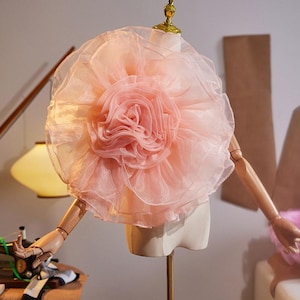 May include: A large, pale pink organza flower accessory featuring multiple layers of sheer, ruffled fabric petals arranged in a circular, voluminous bloom shape, displayed on a wooden mannequin torso with articulated arms and a gold-toned neck cap.