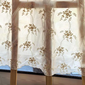May include: Sheer white curtain with embroidered floral design in gold thread. The curtain has a scalloped edge and is hanging in front of a window. The floral pattern is repeated across the fabric.