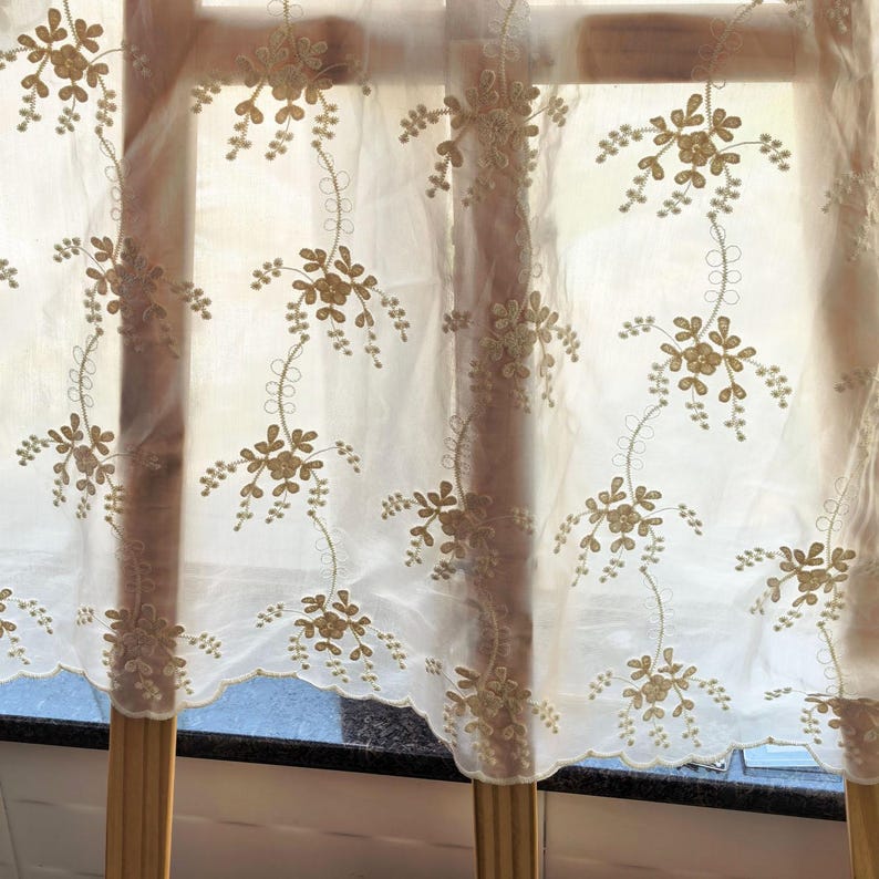 May include: Sheer white curtain with embroidered floral design in gold thread. The curtain has a scalloped edge and is hanging in front of a window. The floral pattern is repeated across the fabric.