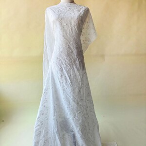 May include: White embroidered fabric draped over a mannequin. The fabric is adorned with delicate floral patterns, creating a textured and elegant appearance. The material appears lightweight and semi-sheer, ideal for dresses or other apparel.