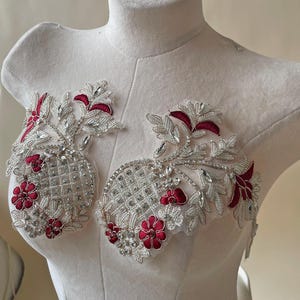 Op de afbeelding: Twee sierlijke, geborduurde applicaties met een bloemenontwerp. De applicaties hebben zilveren draad, heldere strass-steentjes en dieprode bloemen. Het ontwerp wordt getoond op een witte paspop, waarbij de ingewikkelde details van de versieringen worden getoond.