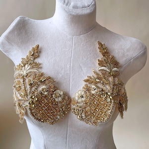 Op de afbeelding: Gouden kralenapplique met bloemen- en bladerenontwerpen. De applique heeft een ruitpatroon en is versierd met kleine, sprankelende kristallen. Het ontwerp wordt getoond op een witte mannequin.