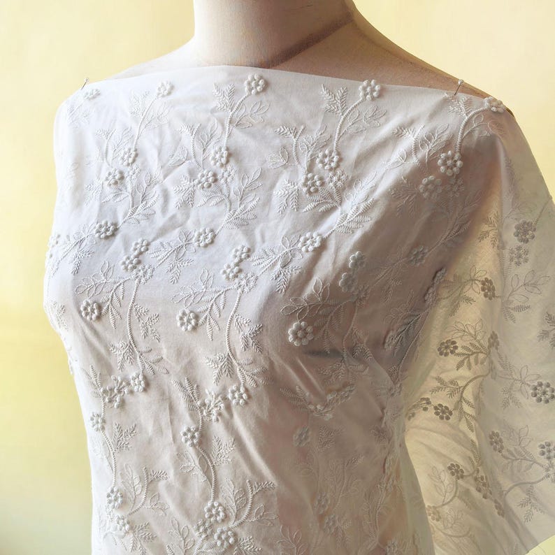 May include: White fabric with an intricate floral embroidery design. The fabric features raised, three-dimensional floral patterns and delicate leaf details. The embroidery is evenly distributed across the fabric, creating a textured and elegant appearance. The fabric is draped over a mannequin.