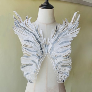 May include: White and gray embroidered angel wings, displayed on a mannequin. The wings feature intricate detailing with gold accents, creating a delicate and ethereal design. The wings are a decorative item, likely for costumes or crafts.