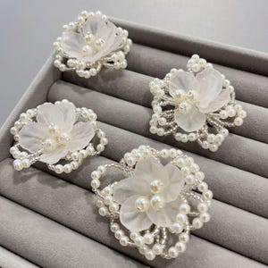 May include: Four white floral brooches with pearl detailing. Each brooch has a layered flower design with a cluster of pearls at the centre, encircled by a beaded frame. The brooches are displayed on a grey surface.
