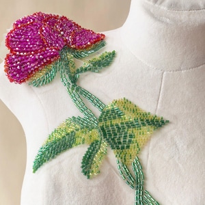 May include: A decorative appliqué featuring a vibrant fuchsia flower head crafted from faceted beads, attached to a stem and leaves made of green and yellow glass bugle beads. The floral design is secured onto a white fabric surface.