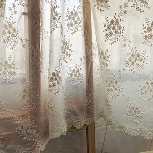 Pu&ograve; includere: Pannelli di tende bianche trasparenti con motivi floreali ricamati in tonalit&agrave; beige e oro. Le tende hanno un bordo in pizzo smerlato lungo il bordo inferiore. La luce filtra attraverso il tessuto, creando un aspetto morbido e arioso.