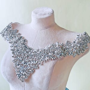 May include: A silver crystal applique, shown on a mannequin. The applique has a V-neck shape and is densely covered with clear crystals of different sizes. The crystals are arranged to create a shimmering, elegant effect, suitable for embellishing clothing.