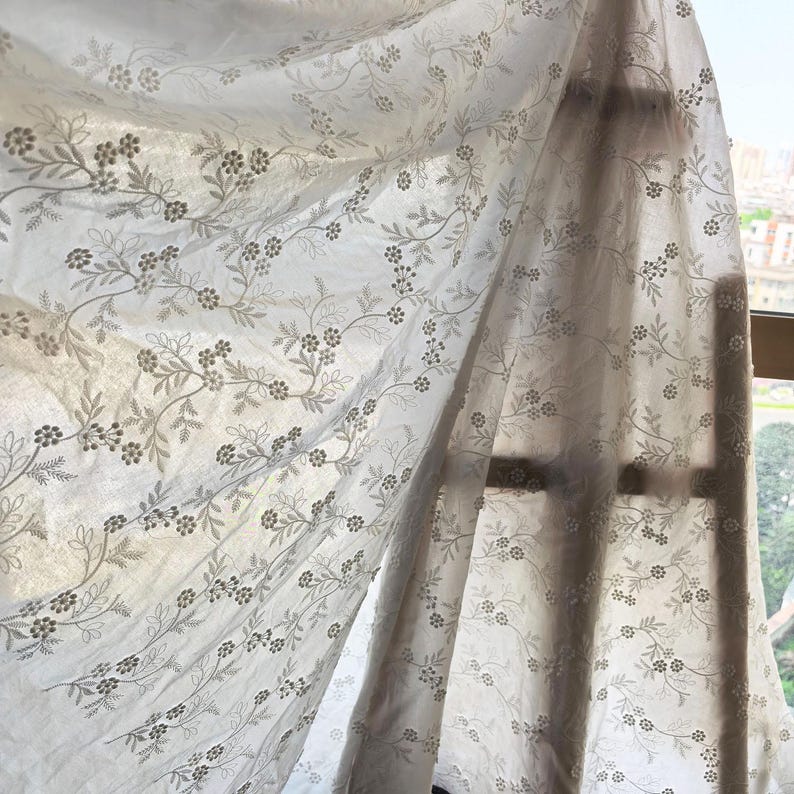 May include: Sheer white curtain with a delicate floral pattern. The design features trailing vines with small flowers and leaves in a muted gray color. The curtain is partially backlit, creating soft shadows and highlighting the fabric's texture.