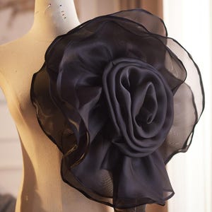 May include: A large, dark charcoal gray organza flower accessory attached to a beige mannequin. The flower features multiple layers of sheer, ruffled fabric petals arranged in a circular, blooming rose shape with a structured, voluminous appearance.