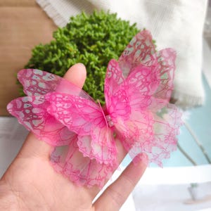 Puede incluir: Decoración de mariposa rosa con intrincados diseños en las alas. La mariposa está hecha de un material transparente y en capas, con un diseño delicado. Las alas tienen un degradado de tonos rosas, y el cuerpo presenta un pequeño adorno decorativo. Es un artículo de artesanía.