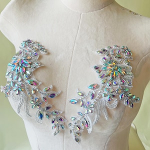 May include: Two floral appliques with iridescent rhinestones and silver beading. The appliques feature a leaf and flower design with a white lace backing. The rhinestones reflect multiple colours, including blue, green, and yellow.