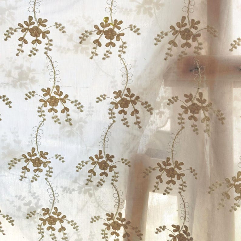 May include: Sheer white curtain with an embroidered floral pattern. The design features gold-colored flowers and delicate stems with small leaves. The curtain is semi-transparent, allowing light to filter through, creating a soft, diffused effect.