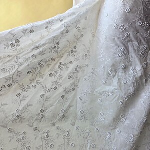 May include: White fabric with delicate floral embroidery. The fabric features a repeating pattern of small flowers and leaves, creating a textured and elegant design. The embroidery is in a subtle, light gray color, adding depth to the fabric.