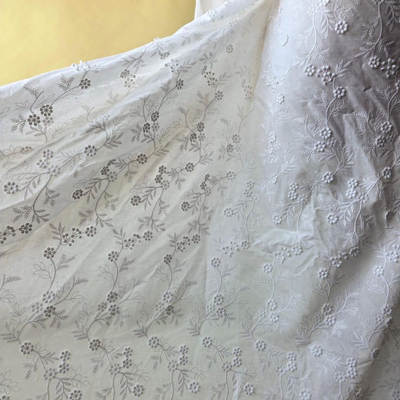May include: White fabric with delicate floral embroidery. The fabric features a repeating pattern of small flowers and leaves, creating a textured and elegant design. The embroidery is in a subtle, light gray color, adding depth to the fabric.