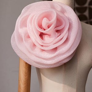 May include: A large, pale pink fabric flower accessory with layered, ruffled petals. The flower has a textured, semi-sheer appearance and is attached to a beige mannequin torso. The petals are arranged in a circular, blooming rose shape.