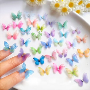 Puede incluir: Una colección de decoraciones de mariposas en miniatura de colores en varios tonos de rosa, azul, verde, amarillo y morado. Cada mariposa tiene un centro de pedrería y alas delicadas y translúcidas. Las mariposas están dispuestas sobre una superficie blanca.