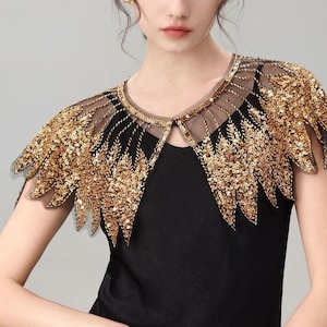 Peut inclure: Haut noir avec une écharpe d'épaule à paillettes dorées. L'écharpe présente un décolleté transparent et des ornements de paillettes dorées dans un motif en forme de plume. Le haut est sans manches et a une encolure arrondie.