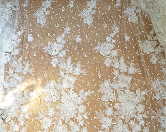 May include: White floral lace fabric with a delicate pattern of embroidered flowers and small, scattered pearl-like beads. The lace is semi-transparent, creating a soft and elegant look, suitable for bridal wear or decorative projects.