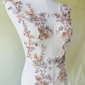 Op de afbeelding: Een sierlijke applicatie in roségoud en kristal, met een bloemmotief. De applicatie is samengesteld uit heldere kristallen en metallic gouden kralen, gerangschikt op een transparante achtergrond. Het ontwerp is symmetrisch en geschikt voor het verfraaien van kleding.