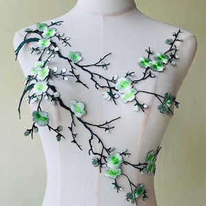 May include: Embroidered floral applique featuring a branch design with green and white flowers. The branches are dark green, and the flowers have a layered appearance. The applique is displayed on a mannequin, showcasing its potential use in fashion or crafts.