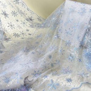 May include: White sheer fabric with a delicate snowflake pattern in shades of blue and silver. The fabric is embellished with sequins and has a scalloped edge, creating a wintery or festive aesthetic.