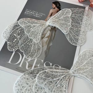 Könnte beinhalten: Zwei weiße, paillettenbesetzte Schmetterlingsflügel-Applikationen mit aufwendigen Details. Die Flügel liegen auf einer Magazinseite mit japanischem Text und dem Wort "DRESS". Die Flügel sind filigran und für ein Kleid oder Kostüm.
