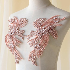 Op de afbeelding: Twee roze-gouden bloemenapplicaties met heldere kristallen accenten. De applicaties zijn versierd met clusters van kristallen en delicate kralenstrengen, waardoor een sprankelend, elegant ontwerp ontstaat. Ideaal voor het verfraaien van bruids- of avondkleding.