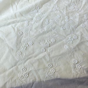 May include: Close-up of white fabric with delicate floral embroidery. The embroidery features detailed stems, leaves, and small flower clusters. The fabric has a soft, slightly wrinkled texture, suggesting it is a lightweight material.