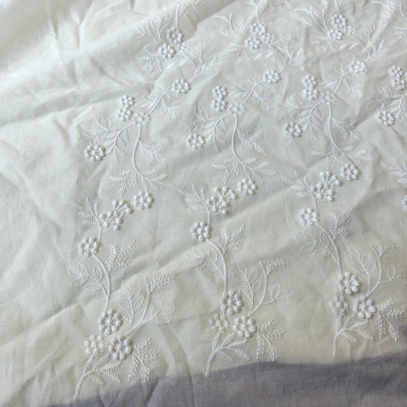 May include: Close-up of white fabric with delicate floral embroidery. The embroidery features detailed stems, leaves, and small flower clusters. The fabric has a soft, slightly wrinkled texture, suggesting it is a lightweight material.