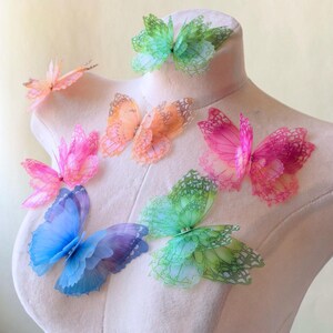Op de afbeelding: Een verzameling kleurrijke vlinderdecoraties in roze, oranje, groen en blauw. De vlinders zijn gemaakt van een dun, delicaat materiaal en zijn op een mannequin geplaatst. De vleugels hebben ingewikkelde patronen.