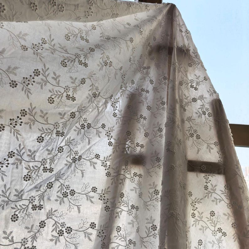 May include: Sheer white fabric with an embroidered floral pattern. The design features delicate gray flowers and leaves on winding stems. The fabric is draped, with sunlight filtering through, creating shadows and highlighting the texture of the embroidery.