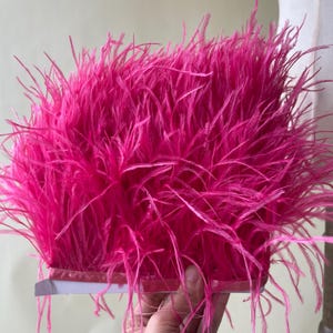 Może przedstawiać: Żywe, intensywnie różowe pióro. Pióra są gęsto upakowane, tworząc puszystą teksturę. Ozdoba jest przymocowana do różowej satynowej wstążki, idealna do rękodzieła i zdobień.