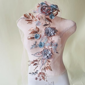 May include: An embroidered floral applique featuring a variety of flowers and leaves in shades of blue, copper, and pink. The applique is embellished with sequins and beads, and is attached to a sheer, light pink fabric.