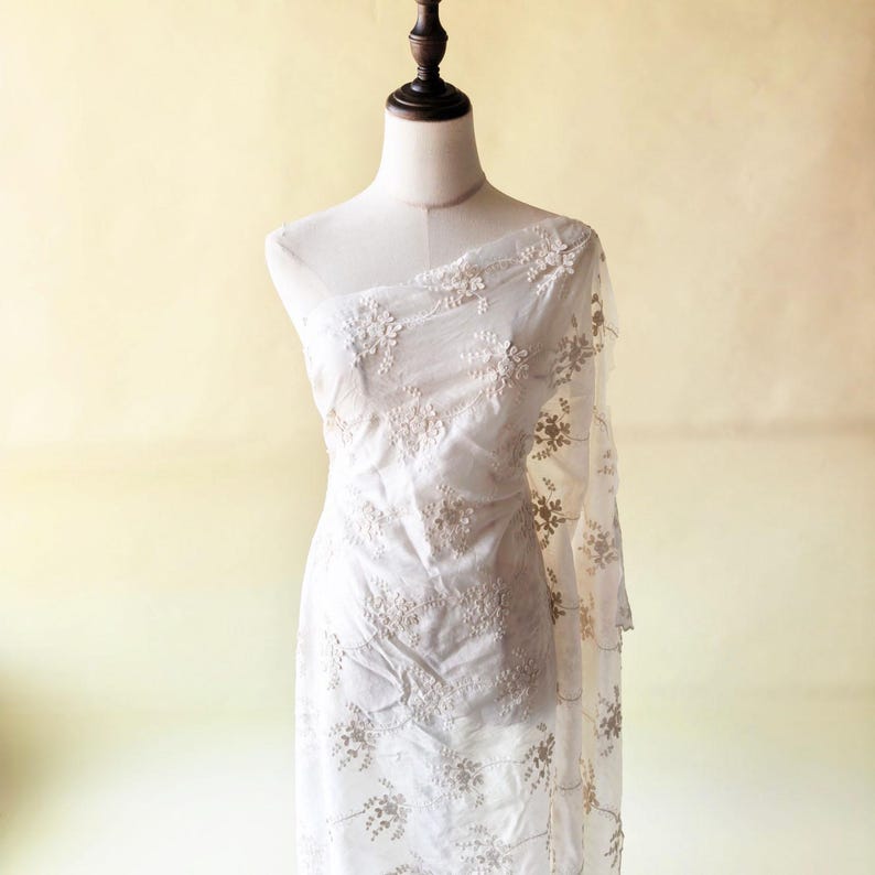 May include: White sheer fabric with floral embroidery draped over a mannequin. The delicate embroidery features a repeating pattern of small flowers and leaves. The fabric is lightweight and appears to be semi-transparent, with a soft, flowing drape.