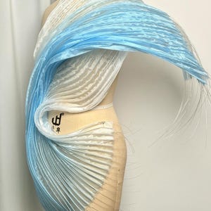 Peut inclure: Un drapé en tissu plissé sur un mannequin, présentant un dégradé passant du blanc au bleu clair. Le matériau a une texture semi-transparente avec de fins filaments s'étendant sur les bords, créant une apparence dynamique et fluide.