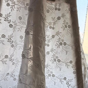 May include: White sheer curtain with embroidered floral and leaf pattern. The design features delicate, small flowers and detailed leaves, creating a soft, textured look. The curtain is partially drawn, allowing sunlight to filter through.