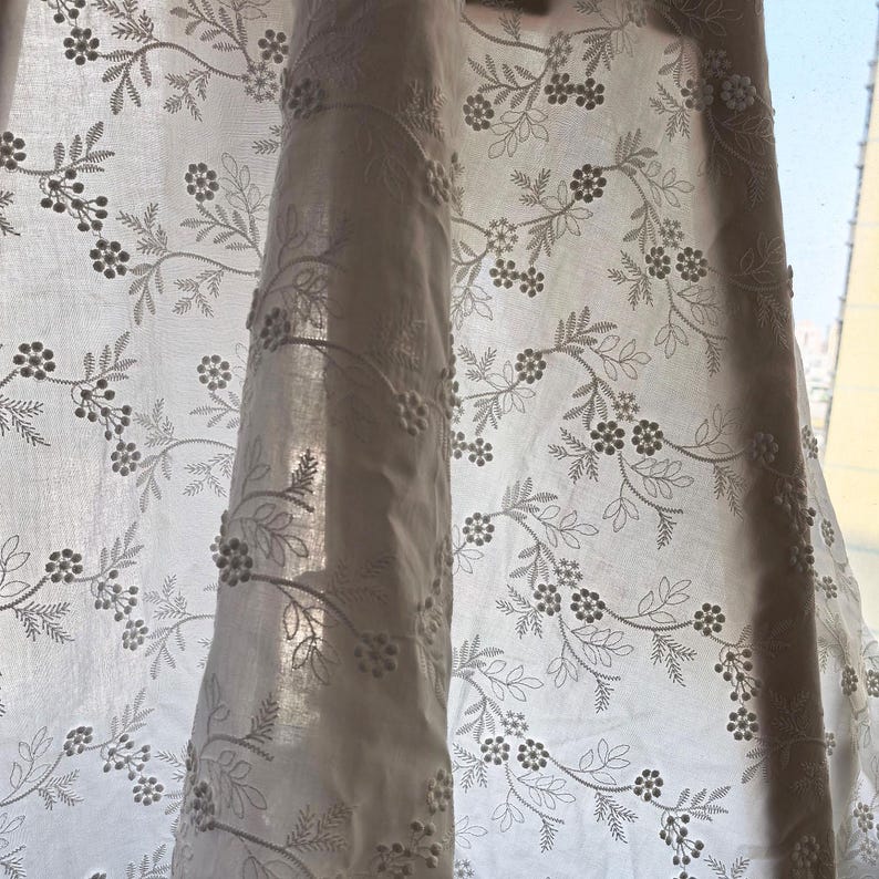 May include: White sheer curtain with embroidered floral and leaf pattern. The design features delicate, small flowers and detailed leaves, creating a soft, textured look. The curtain is partially drawn, allowing sunlight to filter through.