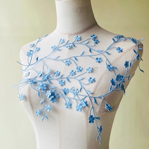 May include: A light blue floral lace applique with embroidered flowers and leaves. The applique has a flowing, vine-like design, suitable for embellishing clothing or crafts. The flowers are a soft, pastel blue, creating a delicate aesthetic.