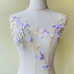 Peut inclure: Applique florale brodée avec des fleurs bleu clair et roses sur des branches jaune-beige. Le design délicat est présenté sur un mannequin, mettant en valeur les détails de l'art textile. Idéal pour embellir des vêtements ou des projets d'artisanat.