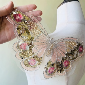 May include: A decorative butterfly applique with pink, gold, and pearl embellishments. The butterfly features sequins, beads, and floral accents, with a silver-colored wire outline. The wings are a soft pink color, and the applique is on a sheer mesh backing.