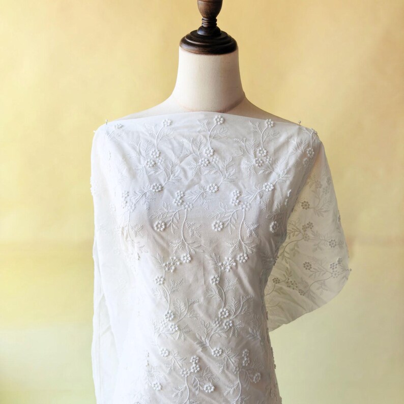 May include: White embroidered fabric with a floral pattern. The fabric is draped over a mannequin, showcasing the delicate, detailed design. The embroidery features small, raised floral elements and leafy vines, creating a textured effect.