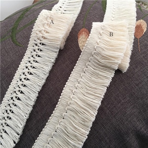 Puede incluir: Se muestran dos adornos de flecos blanquecinos. El adorno A presenta un diseño geométrico calado, mientras que el adorno B tiene un fleco más denso. Ambos adornos están hechos de un material tejido suave, ideal para proyectos de manualidades.