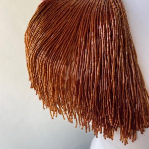 May include: Close-up of a shoulder detail featuring a cascade of brown beaded fringe. The beads are small, cylindrical, and a warm, earthy tone. The fringe hangs in vertical lines, creating a textured, layered effect. The background is a soft, neutral color.